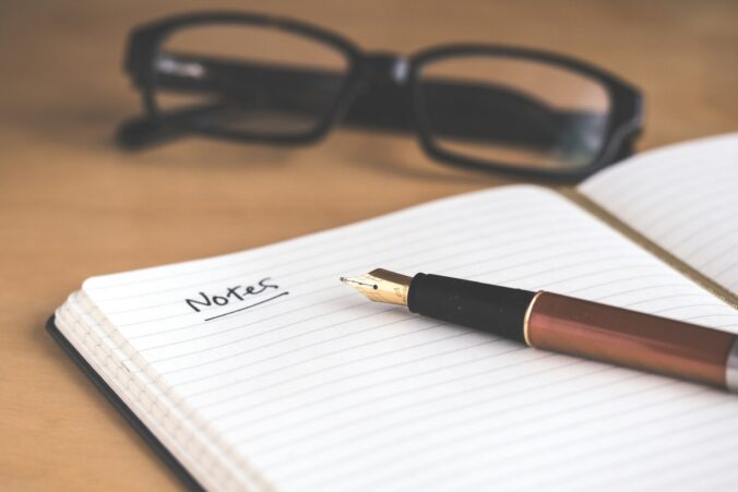 Image is of an open notebook on a wood table. There's a fountain pen lying across the page and the word "Notes" written on an otherwise blank page. A pair of black reading glasses sit on the table above the notebook.